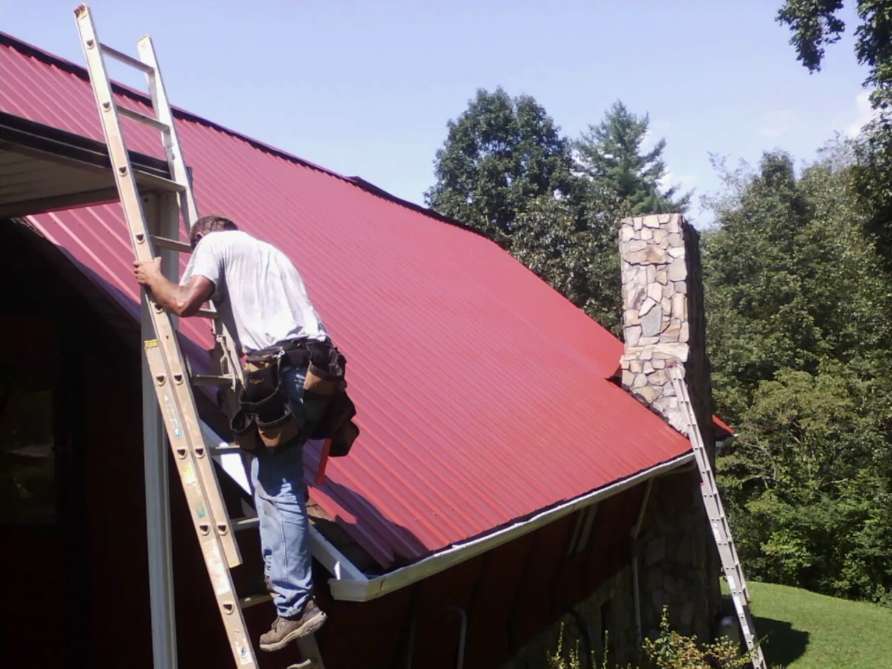 Roofer on a ladder beside a red metal roof and stone chimney — Soffit & Fascia Repair by Roofing Brothers in Hickory Flat